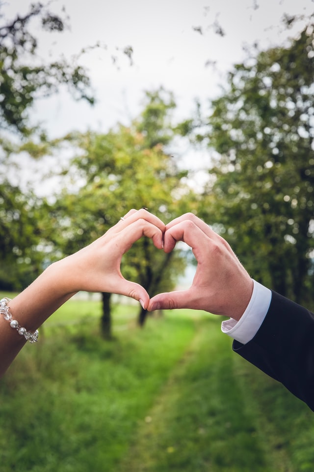 Bride and groom each have one hand forming a heart against a grassy field