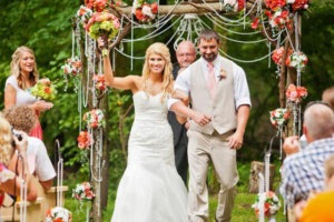 Bride and groom walk hand-in-hand under a rustic wooden wedding altar adorned with florals, greenery, and hanging crystal strands during an outdoor ceremony.