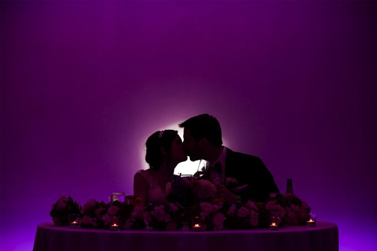 kissing wedding couple seated at the sweetheart table with purple uplighting at a wedding venue in in Lansdale PA 