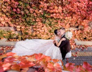 A bride and groom embrace and smile in front of a vibrant wall of red, orange, and green fall leaves, with the bride’s gown spread out around them